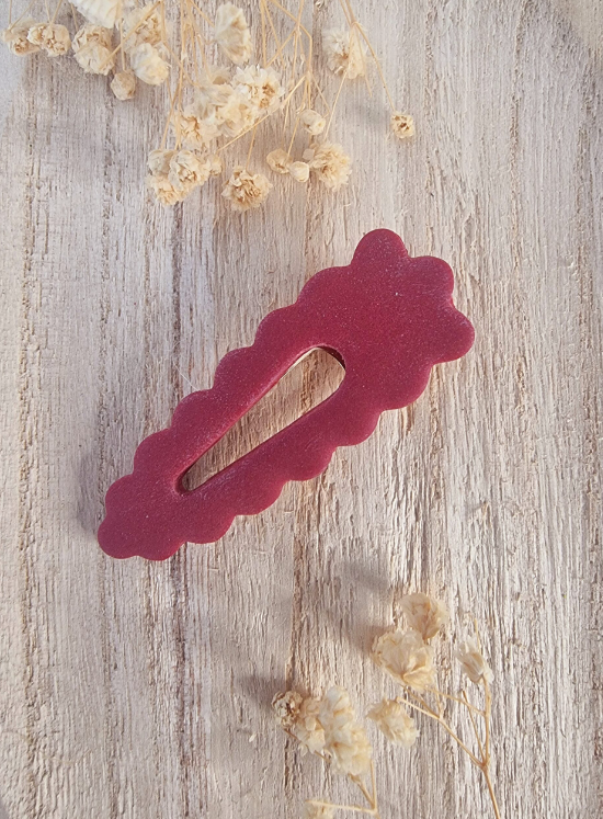 Barrette de forme triangulaire posée sur un support en bois entouré de fleurs. Faite main en argile polymère et en acier inoxydable. Ton bordeaux. Modèle Barrette Fleurie, style classique.