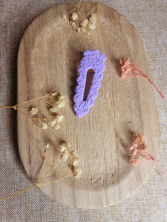 Barrette de forme triangulaire posée sur un support en bois entouré de fleurs. Faite main en argile polymère et en acier inoxydable. Ton mauve. Modèle Barrette Fleurie, style classique et floral.