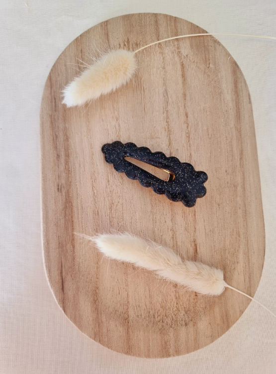 Barrette de forme triangulaire posée sur un petit plateau en bois. Faite main en argile polymère et en acier inoxydable. Ton noir. Modèle Barrette Fleurie, style classique.