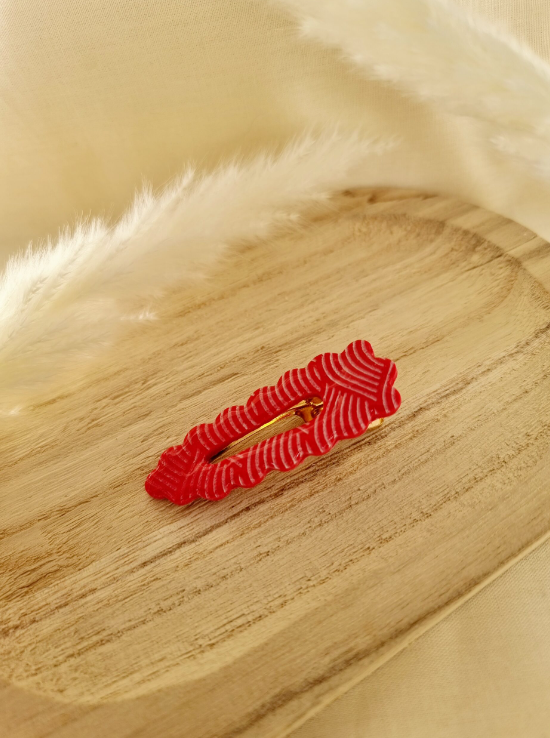 Barrette de forme triangulaire posée sur un support en bois entouré de fleurs. Faite main en argile polymère et en acier inoxydable. Tons rouge et blanc. Modèle Barrette Fleurie, style classique.