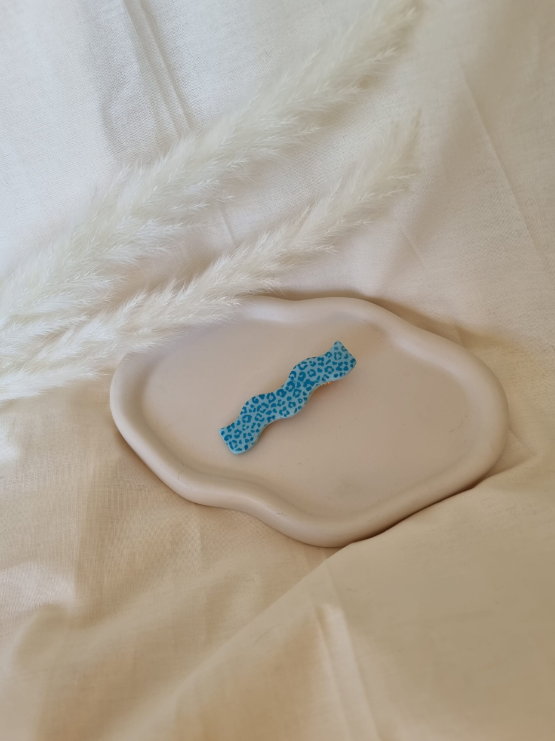Barrette en forme de vague posée sur un petit plateau en bois. Faite main en argile polymère et en acier inoxydable. Ton bleu. Modèle Barrette Vague, style classique.