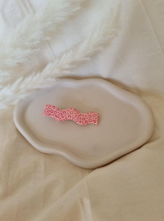 Barrette en forme de vague posée sur un petit plateau en céramique blanc. Faite main en argile polymère et en acier inoxydable. Tons roses. Modèle Barrette Vague, style classique.