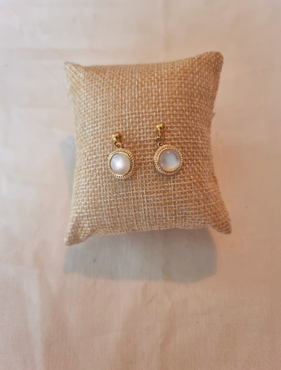 Boucles d'oreilles en forme de sphère. Faites main en nacre et en acier inoxydable. Posées sur un petit coussin en toile. Vue rapprochée. Tons blanc et doré. Modèle Anthéa, style classique.