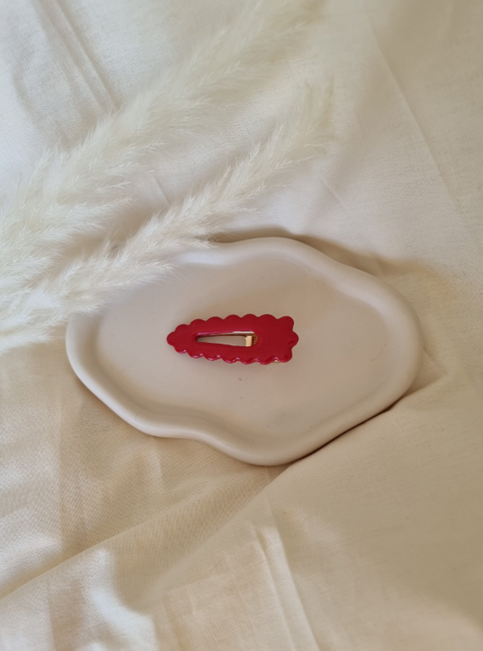 Barrette de forme triangulaire posée sur un support en céramique blanc entouré de fleurs. Faite main en argile polymère et en acier inoxydable. Ton rouge. Modèle Barrette Fleurie, style classique.