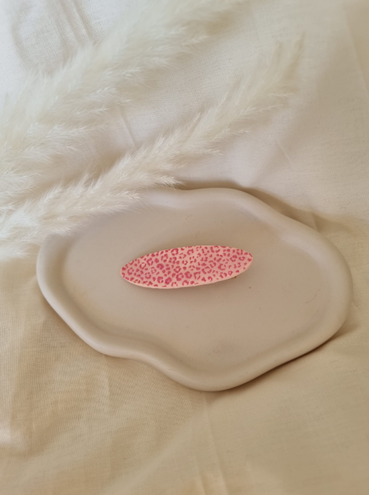 Barrette de forme ovale posée sur un support en bois. Faite main en argile polymère et en acier inoxydable. Ton léopard rose. Modèle Barrette Ovale, style classique.