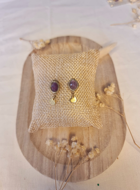 Boucles d'oreilles en forme d'oeuf, avec un petit coeur accroché. Faites main en améthyste et en acier inoxydable. Posées sur un petit coussin en tissu entouré de fleurs. Tons mauve et doré. Modèle Anémone, style classique et romantique.